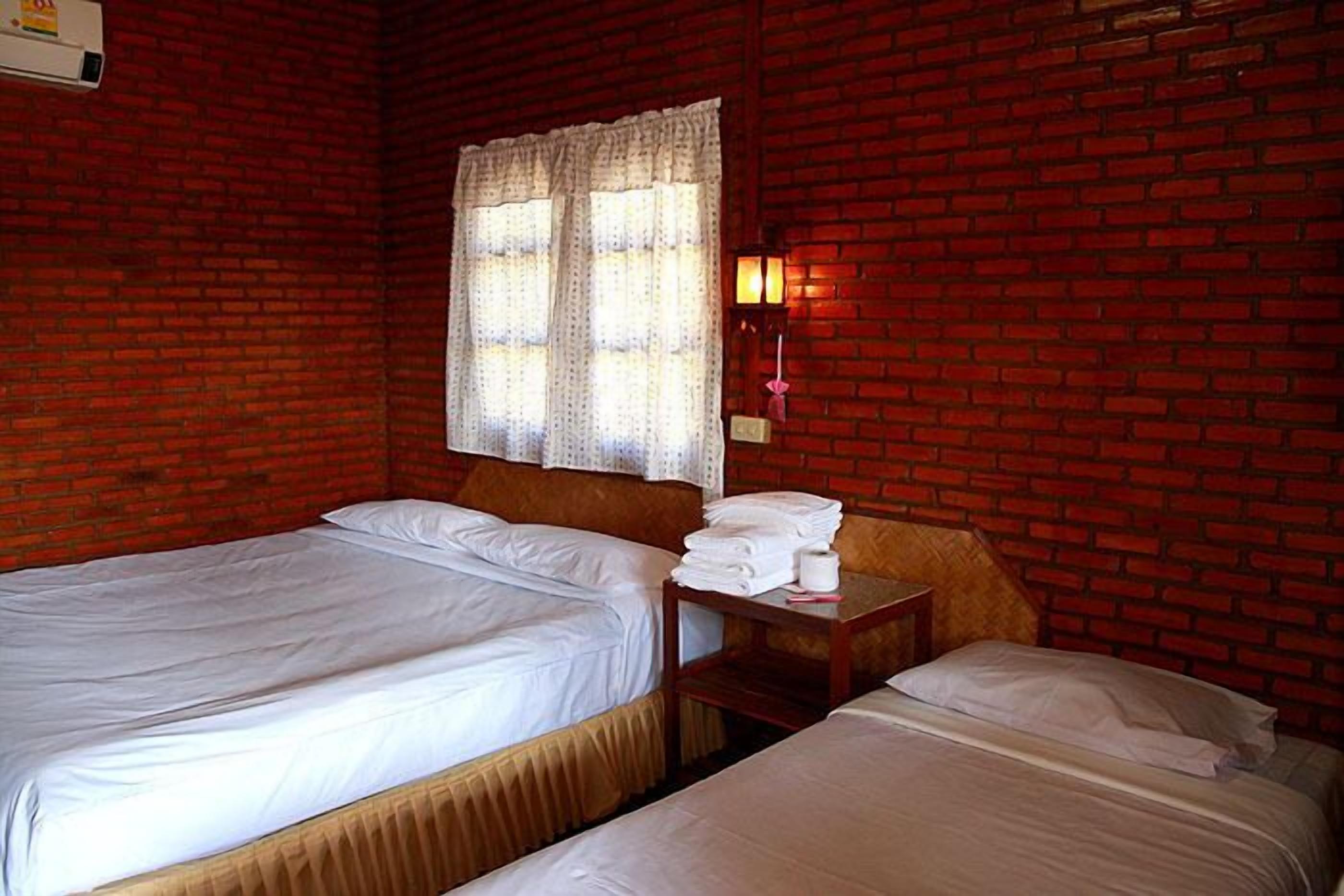 A room with two beds, a window with curtains, a wall-mounted air conditioner, and a nightstand with towels.