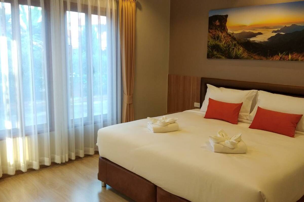 A hotel room with a large bed, white linens, red pillows, and a painting of a landscape on the wall.