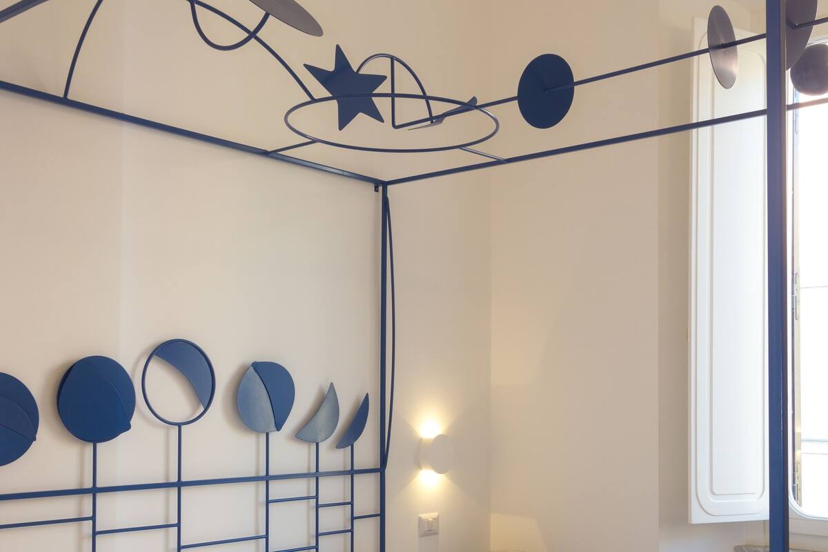 A bed with a metal frame and a canopy, a bedside table with a lamp, and a window with a view of the outside.