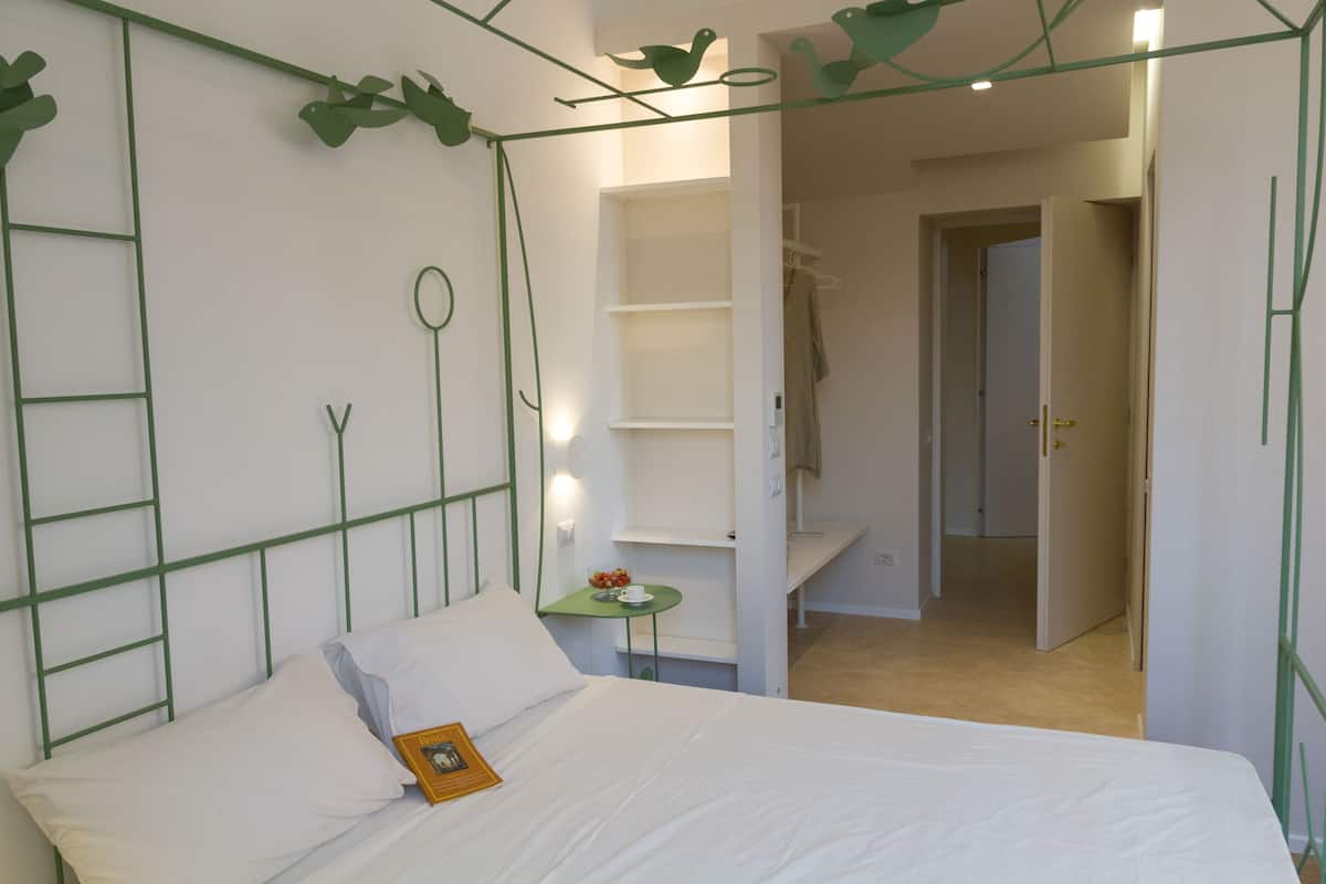 A bedroom with a bed, a small bedside table, a wall-mounted shelf, and a door leading to another room.