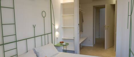A bedroom with a bed, a small bedside table, a wall-mounted shelf, and a door leading to another room.