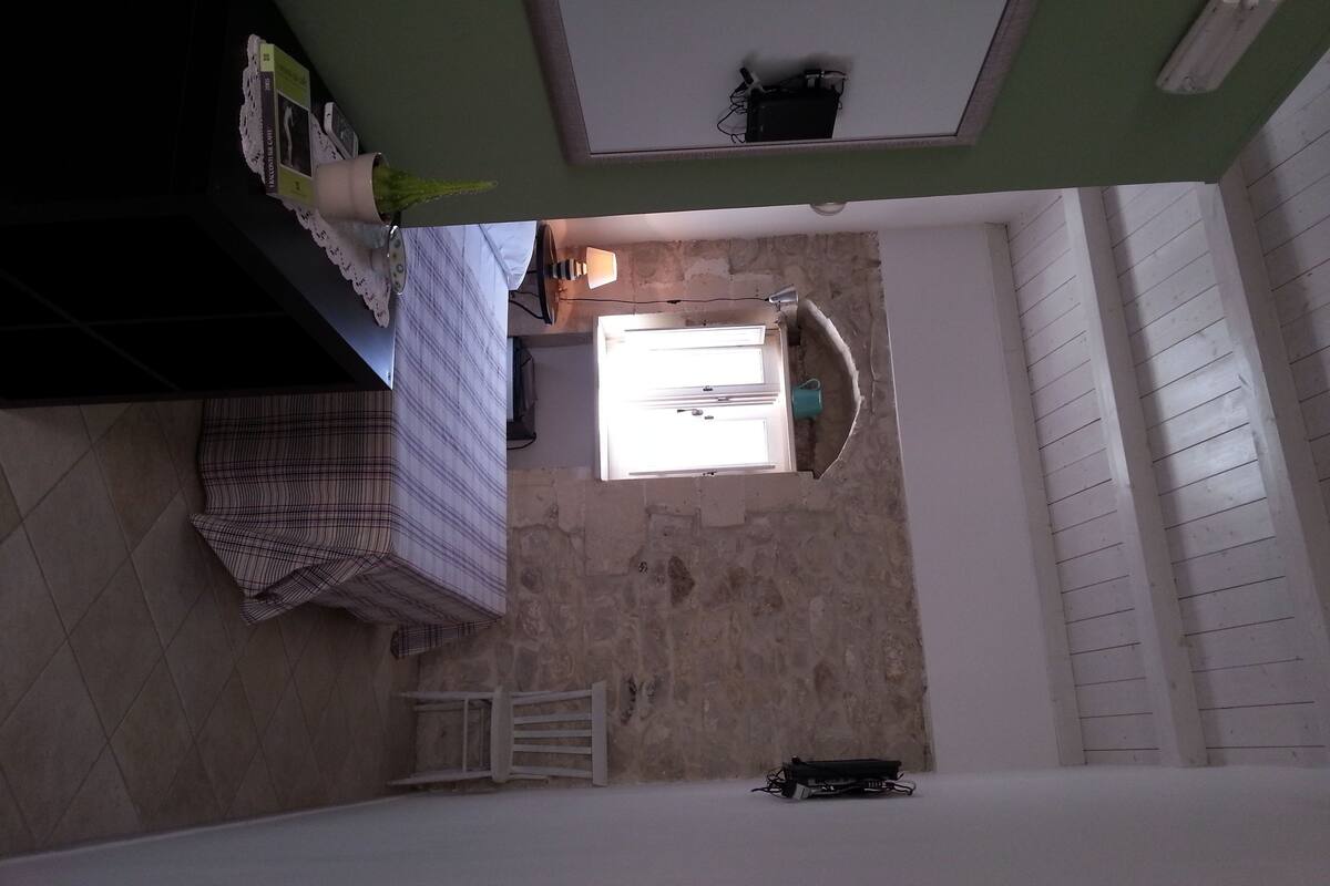 Une pièce avec un sol carrelé, un lit recouvert d’une couverture à rayures, une table de chevet avec une lampe, une petite plante en pot et une fenêtre donnant sur l’extérieur.