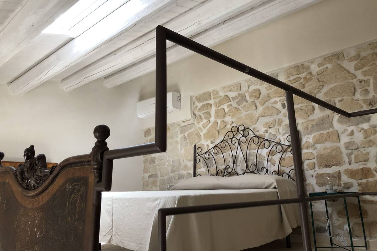 Une chambre avec un mur en pierre, un lit avec un cadre en métal, une tête de lit en bois et un puits de lumière.