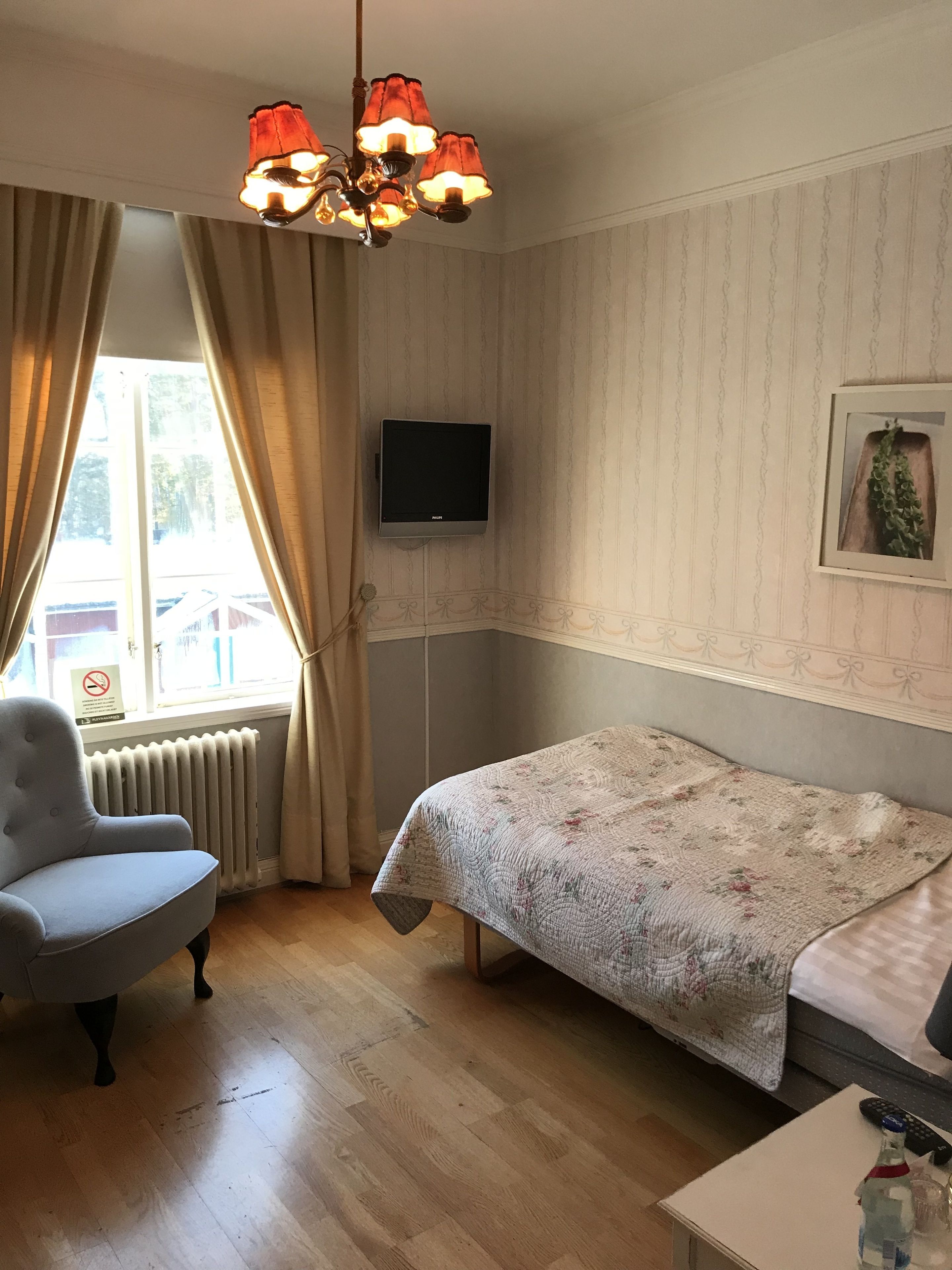 A room with a bed, a chair, a television on the wall, a radiator, and a window with curtains.