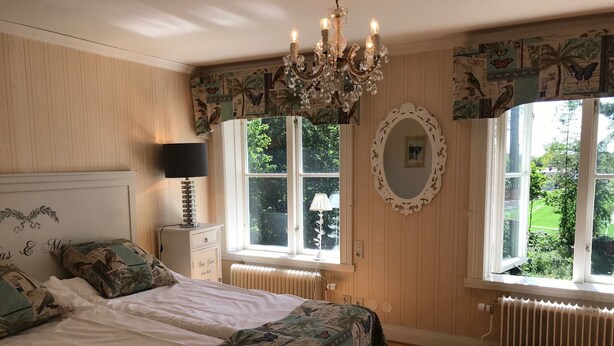 A bedroom with a bed, a chandelier, a window with a view of greenery, a radiator, and a patterned bedspread. - Hotell Plevnagården (Malmkoping)