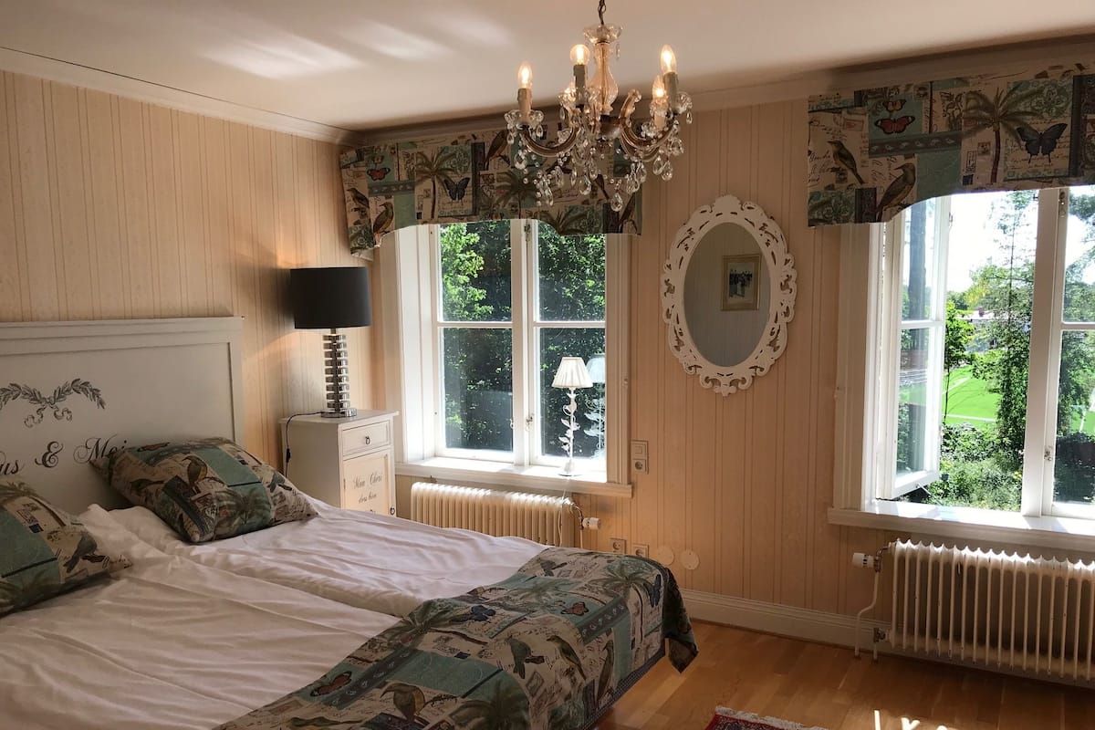 A bedroom with a bed, a chandelier, a window with a view of greenery, a radiator, and a patterned bedspread.