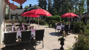 An outdoor patio with tables and chairs, red umbrellas, and a forested background. - Hotell Plevnagården (Malmkoping)