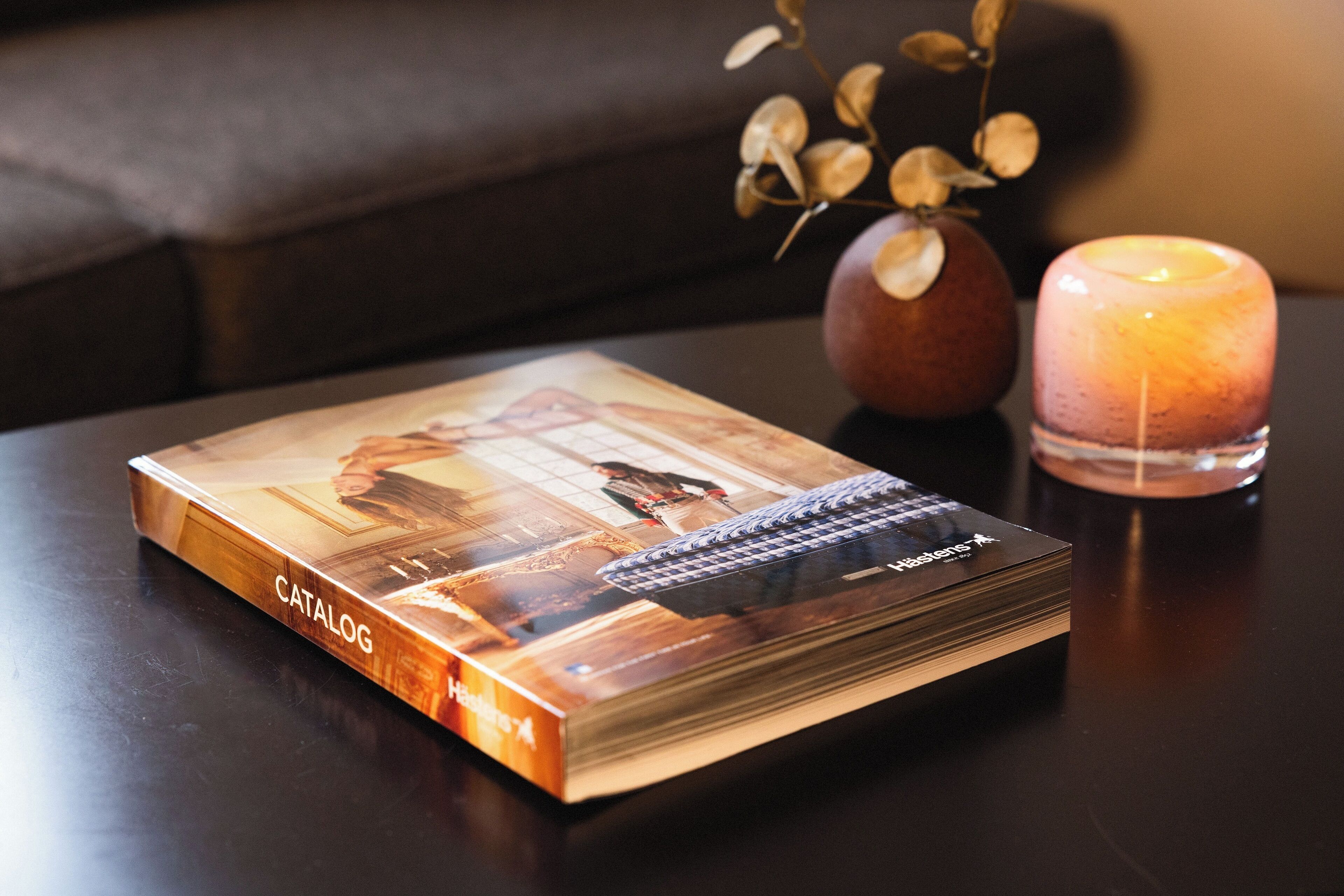 Un libro titulado 'CATALOG' sobre una mesa con una vela y un florero con hojas secas.