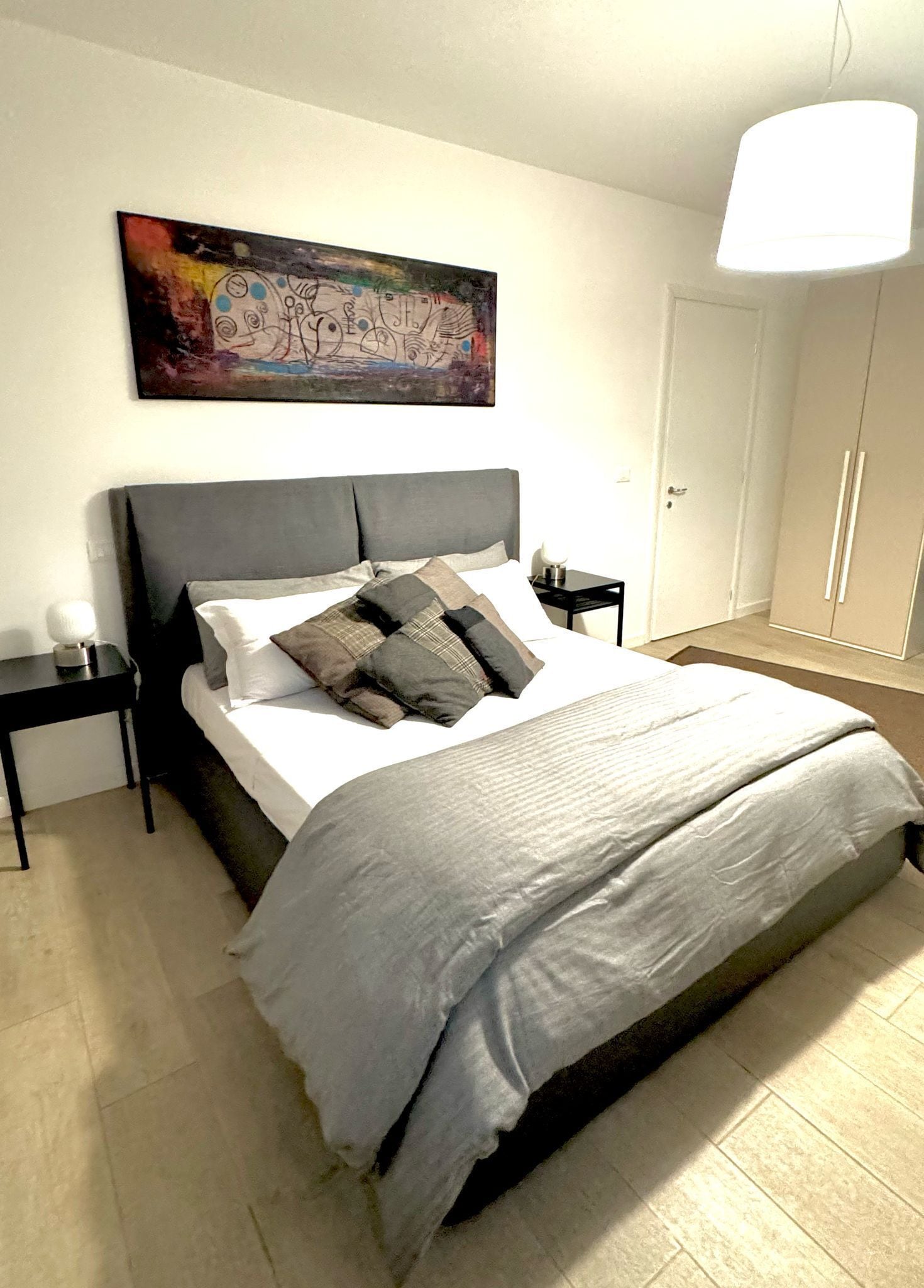 Een netjes opgemaakt bed met een grijs hoofdbord, een nachtkastje met een lamp en een schilderij aan de muur.