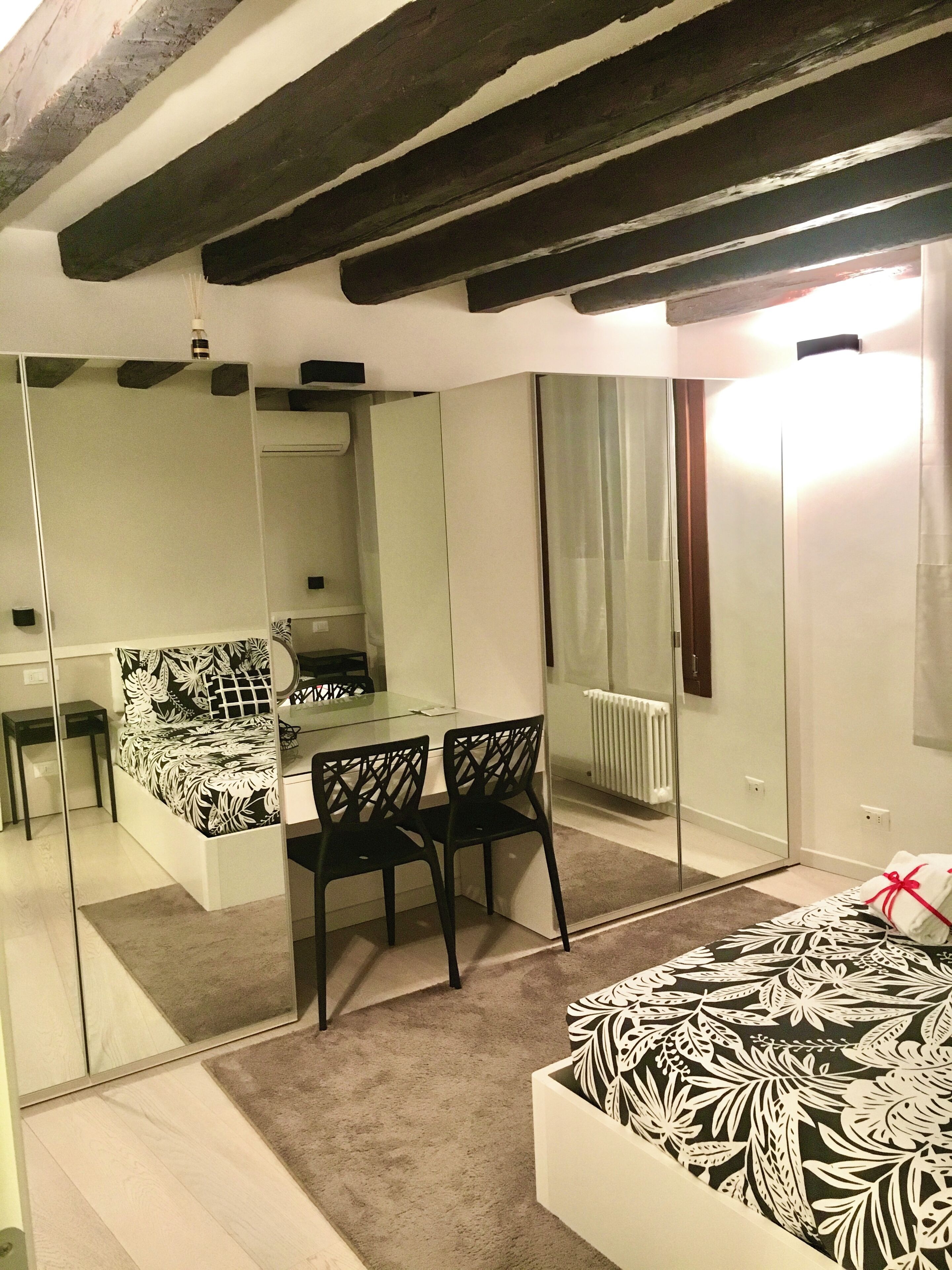 A modern bedroom with a large mirror, a bed with a floral bedspread, a small desk with a chair, and a radiator.