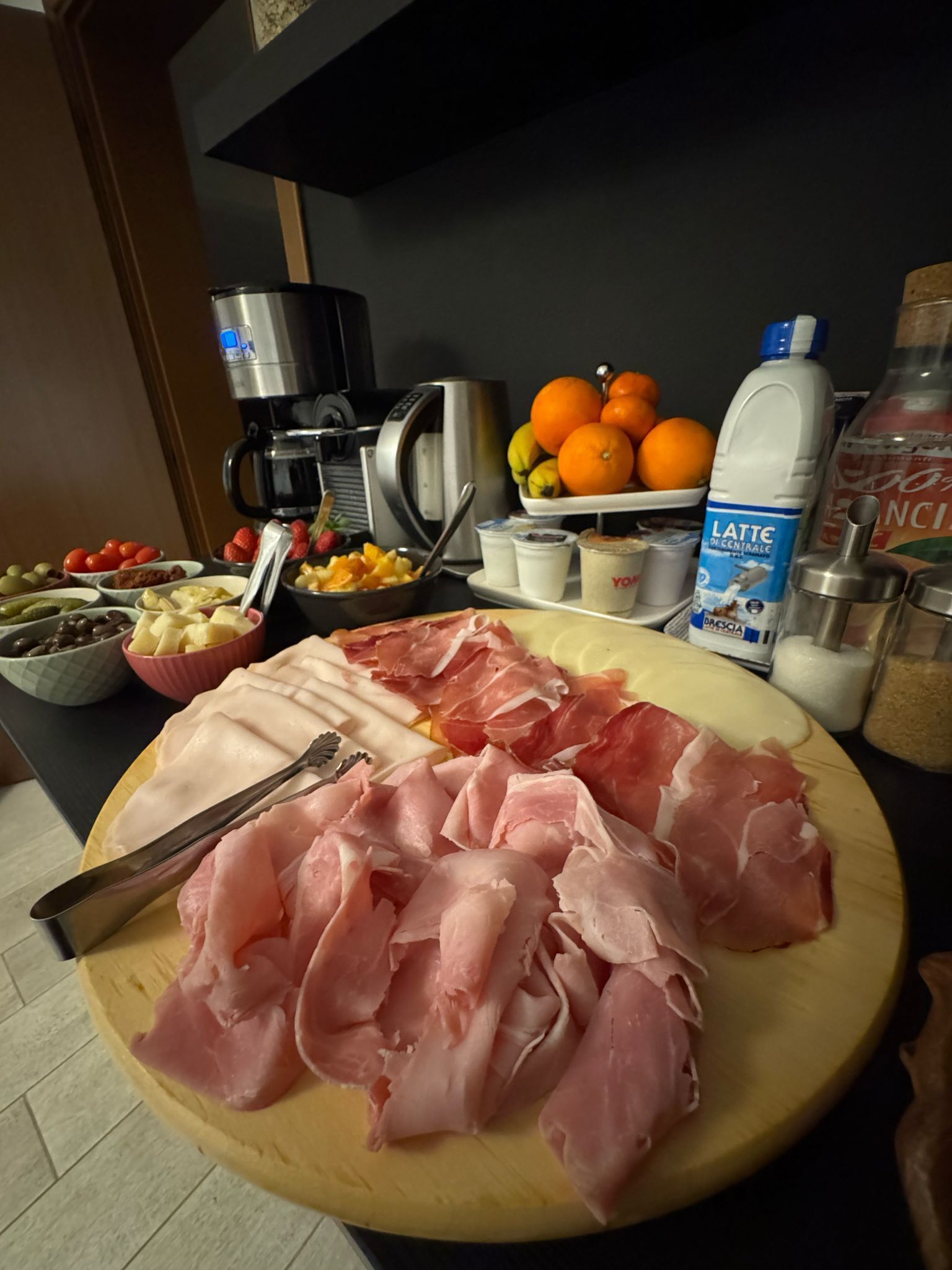 A wooden cutting board with various meats and a knife, a coffee maker, a fruit bowl, and a milk carton on a kitchen counter.