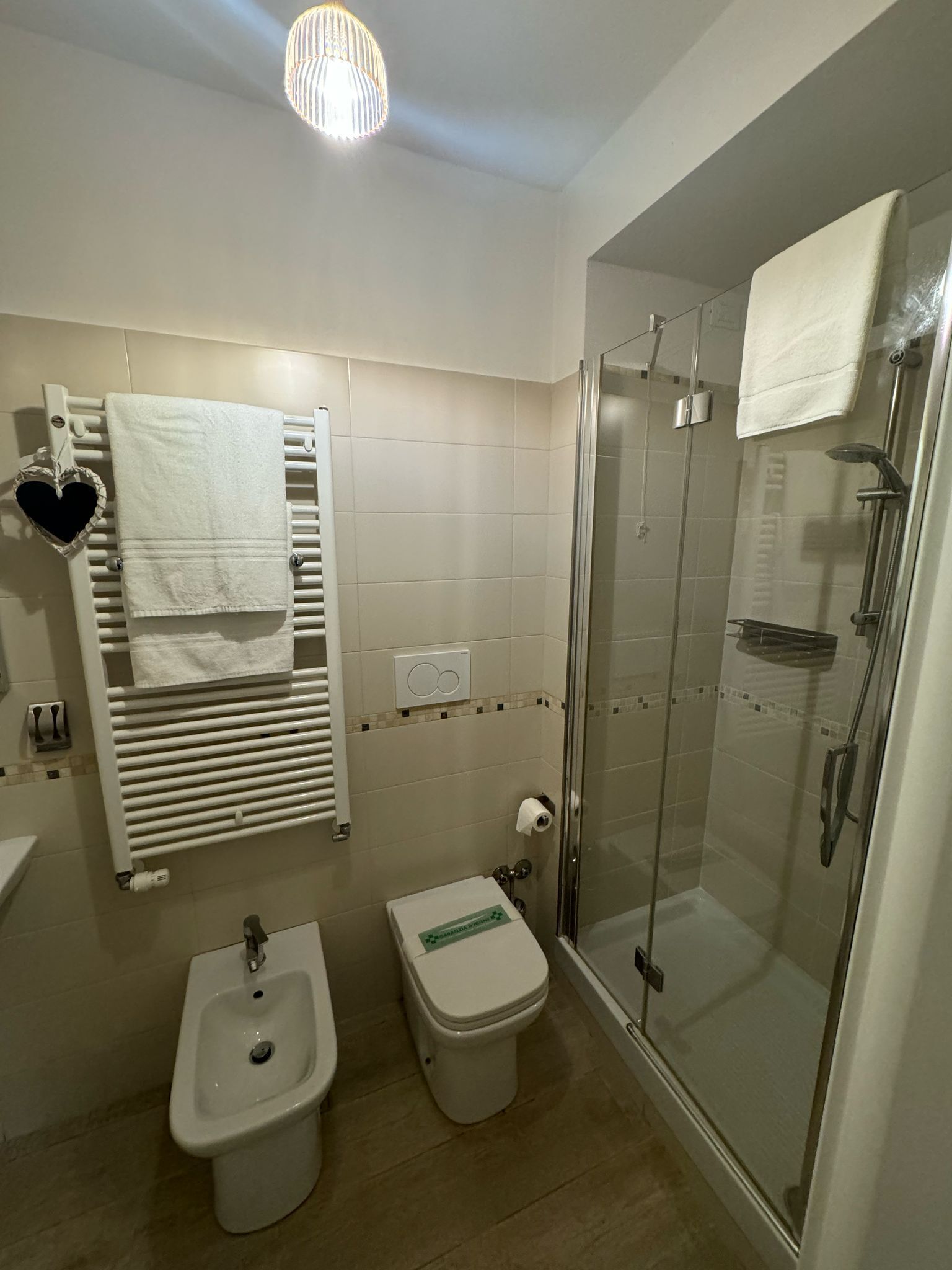 Un baño moderno con ducha de vidrio, un bidé, un inodoro y un perchero con un adorno con forma de corazón.