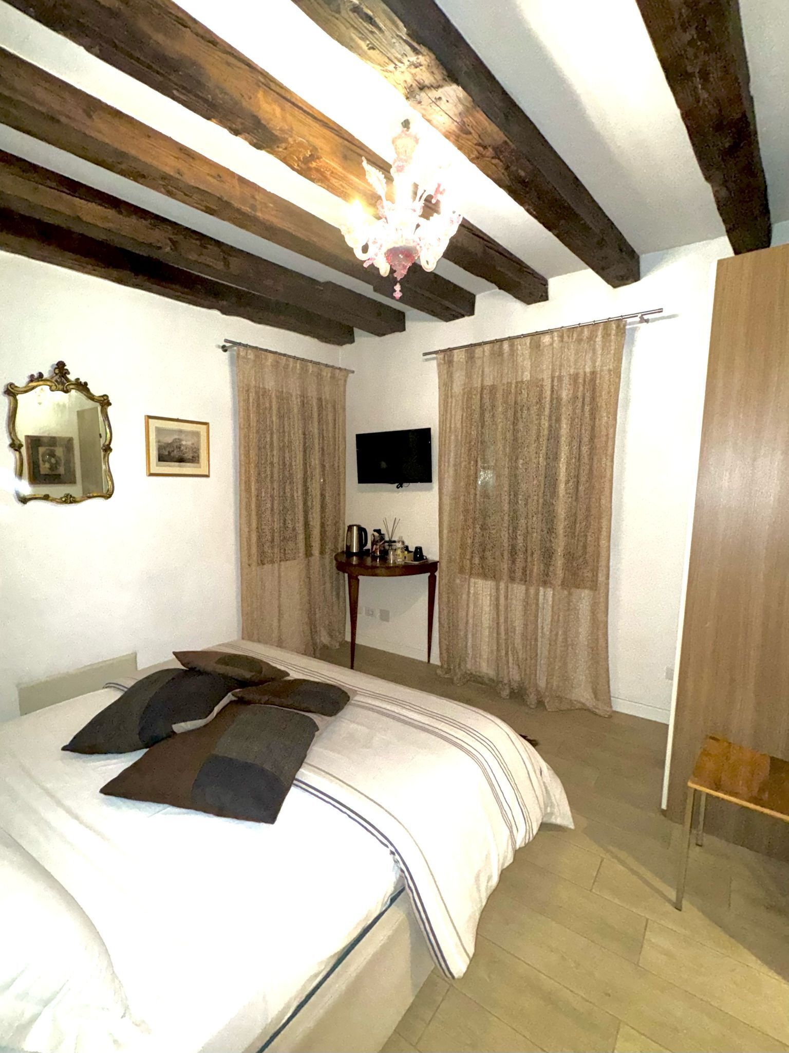 A bedroom with a bed, wooden beams, a television, curtains, a small table, and a mirror.
