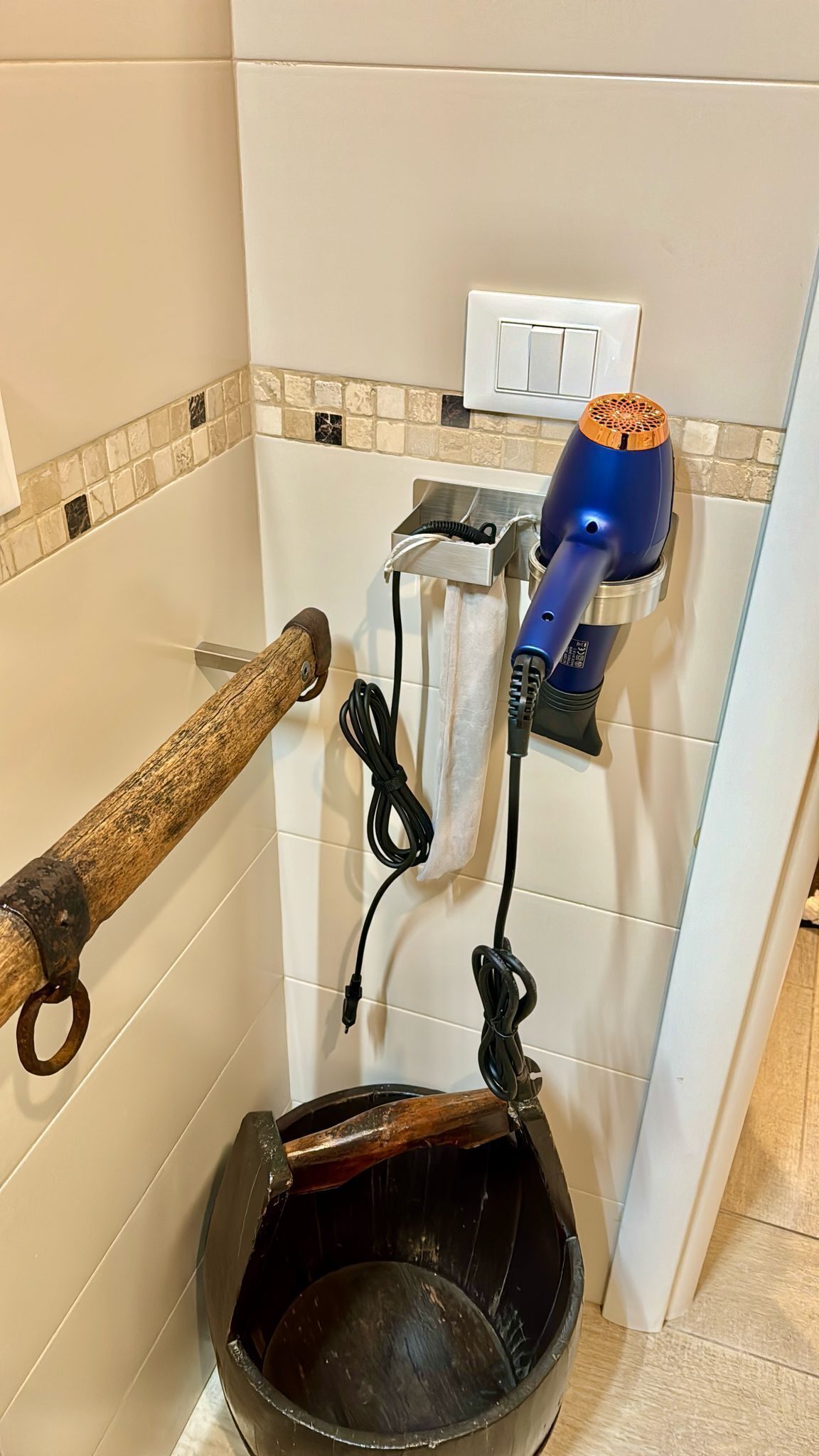 A bathroom with a hairdryer mounted on the wall, a wooden towel rack, and a black bucket.
