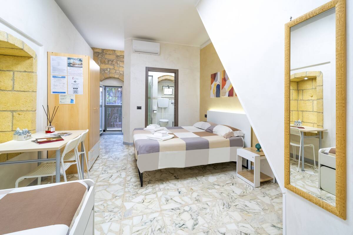 A modern hotel room with a bed, a desk, a chair, a small table, and a mirror.