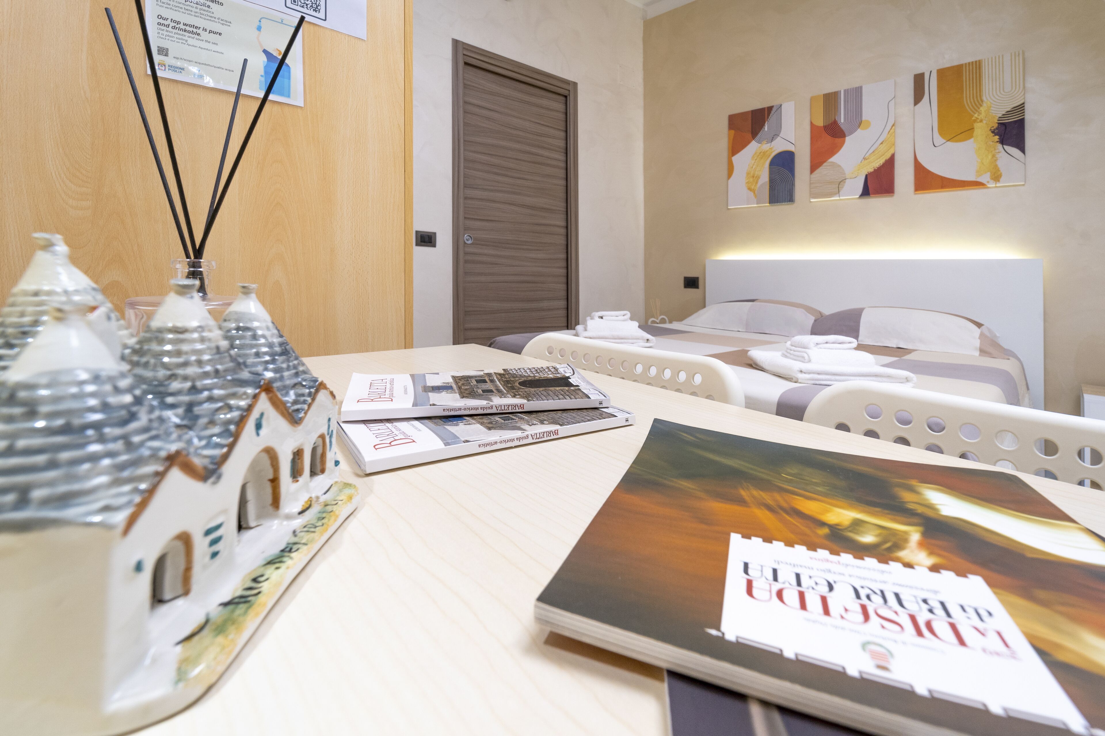 A hotel room with a bed, a desk with magazines, a small model house, and abstract wall art.