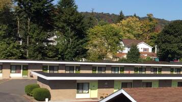A motel with a row of rooms, each with a window and a door. The building is surrounded by trees and a hill in the background.