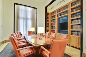 Uma sala de conferências com uma mesa de vidro, cadeiras de couro, uma janela grande com cortinas e uma estante com livros e uma TV.