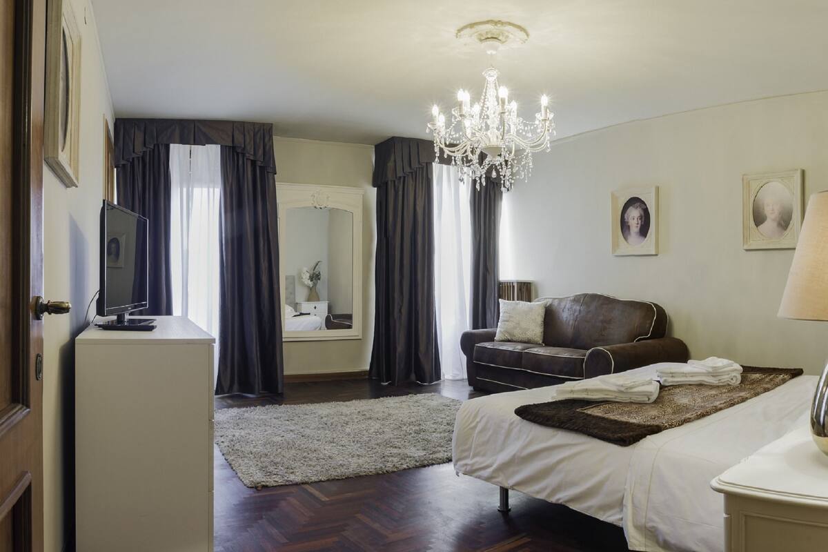 Una habitación de hotel con una cama, un escritorio con un ordenador, una araña, un sofá y una lámpara.