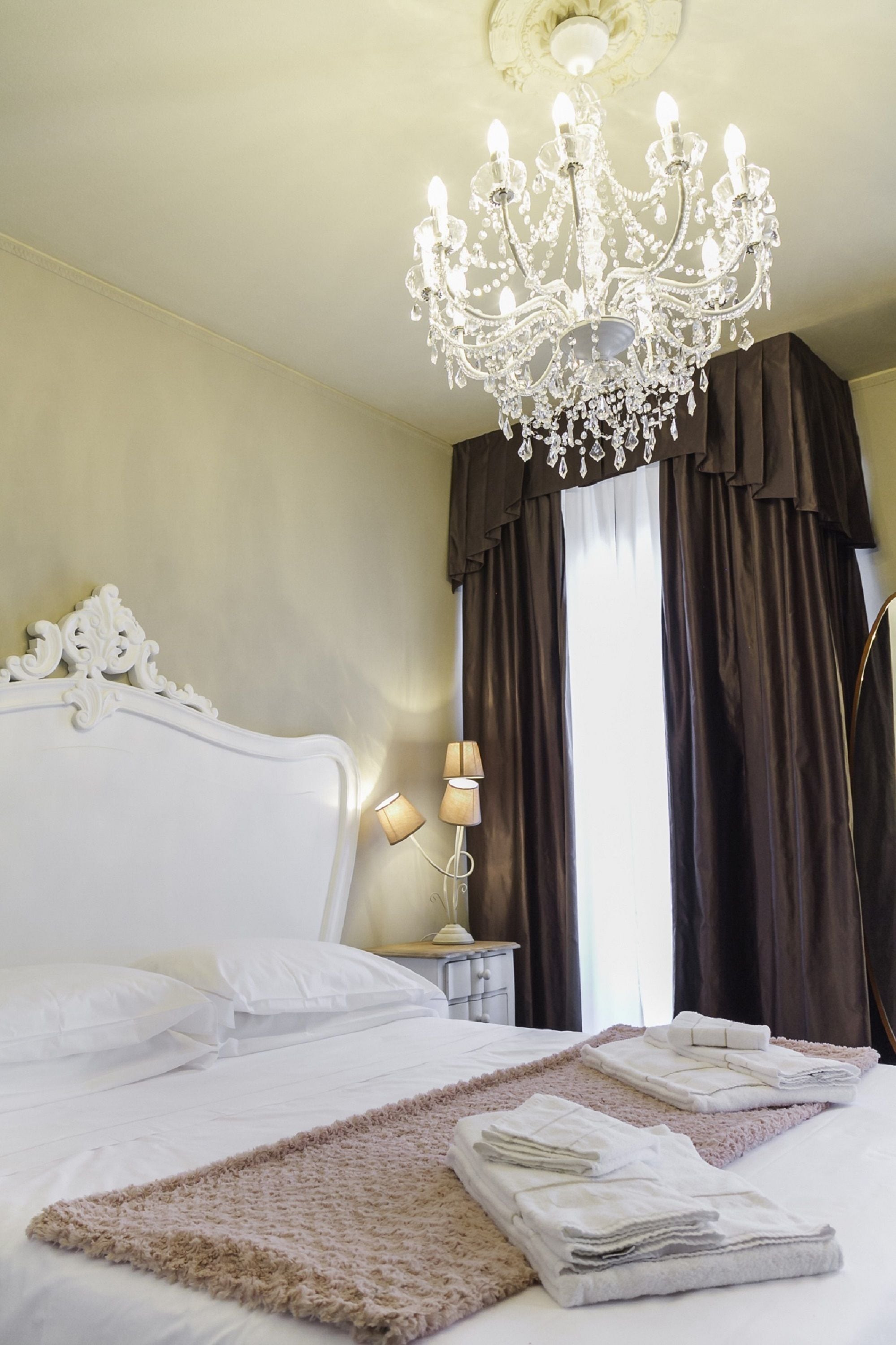 A hotel room with a large bed, a chandelier, a nightstand with a lamp, and a window with curtains.