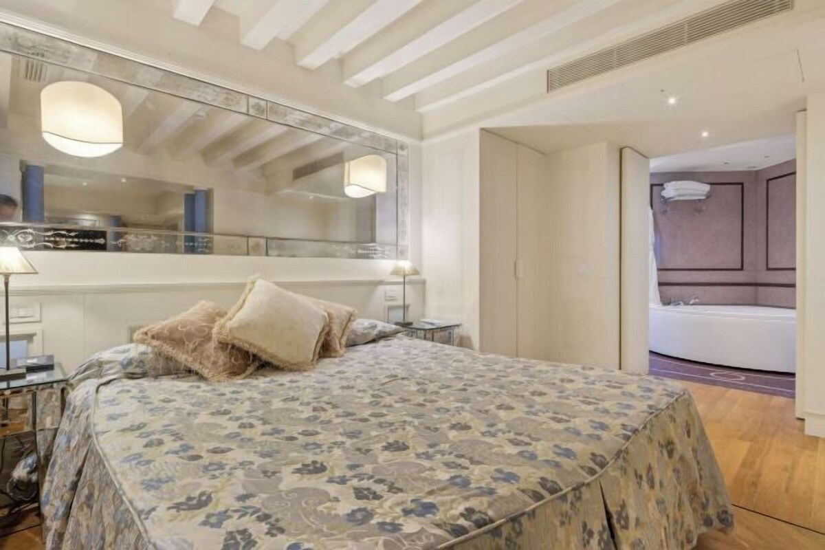 A bedroom with a large bed, a mirror, bedside tables, and a bathroom visible through an open door.