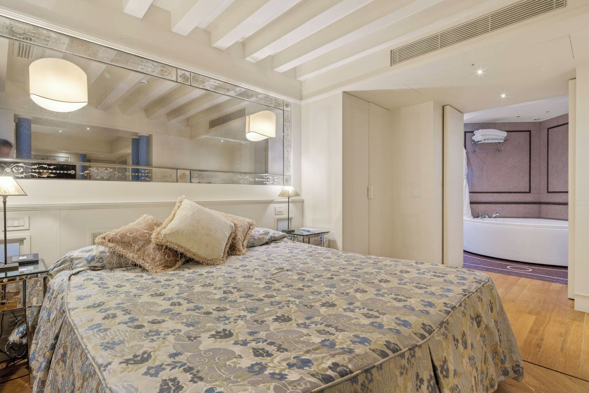 Quarto com uma cama grande, mesa de cabeceira, espelho e banheiro visível por uma porta aberta.