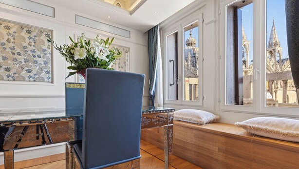 Une pièce avec un bureau en bois, une chaise bleue, un vase de fleurs et une vue sur un bâtiment historique, visible à travers de grandes fenêtres.