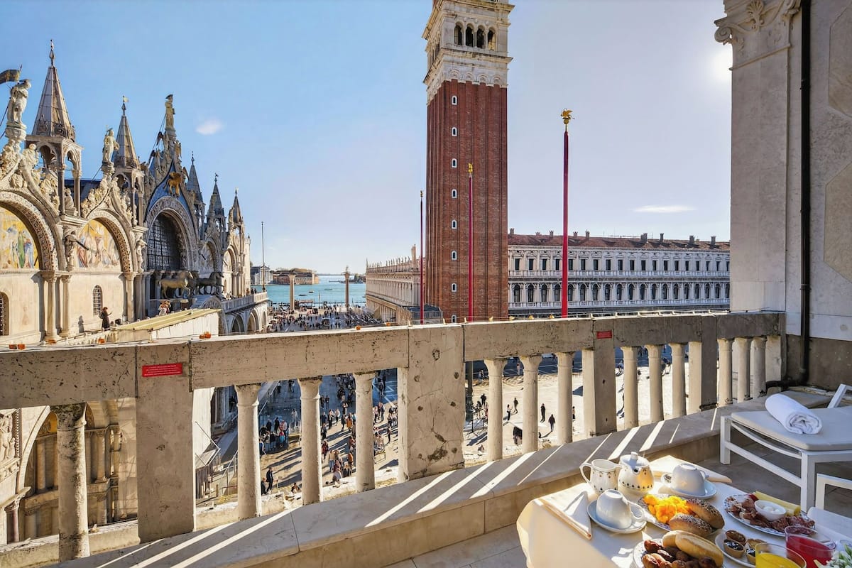 Un balcon avec une table dressée pour le petit-déjeuner, offrant une vue sur un paysage urbain historique, avec des bâtiments richement décorés et un imposant beffroi.