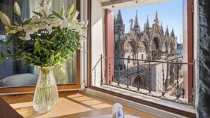 A room with a view of a historic building with ornate architecture, a balcony with a wrought iron railing, and a vase with white flowers on a wooden table.