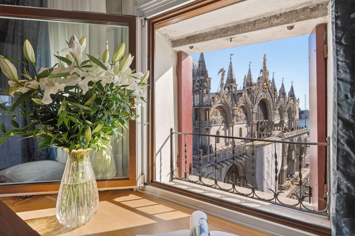 Une pièce offrant une vue sur un bâtiment historique à l’architecture riche et ornée, un balcon doté d’une balustrade en fer forgé et un vase rempli de fleurs blanches posé sur une table en bois.