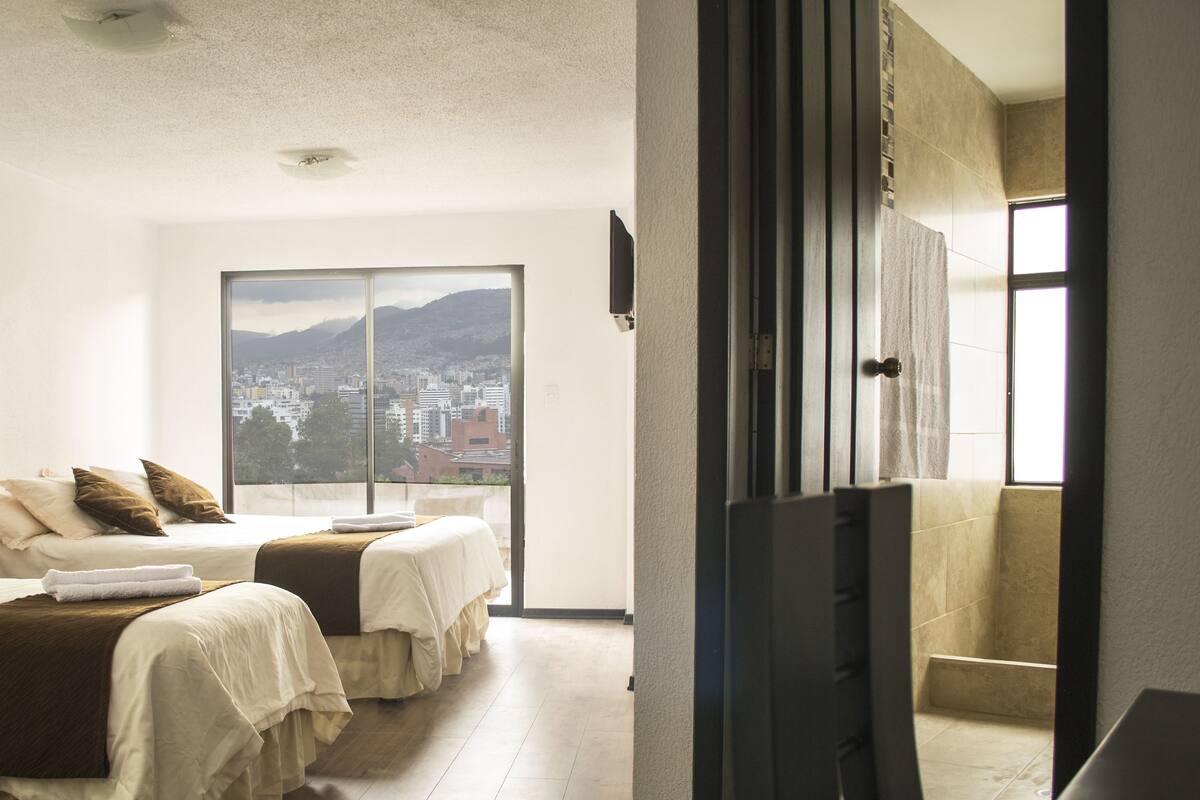 A hotel room with two beds, a large window with a city view, and a flat-screen TV mounted on the wall.
