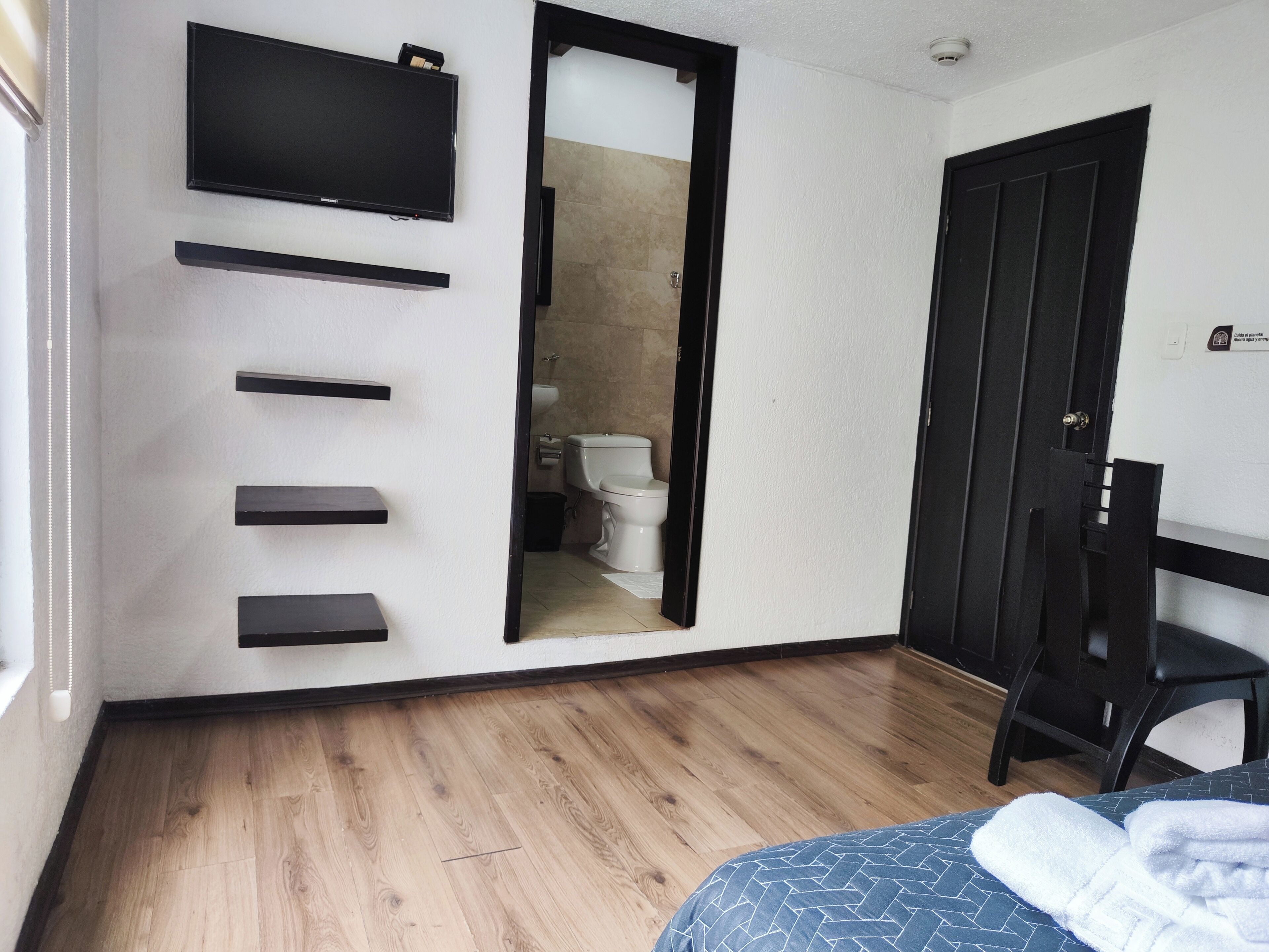 A hotel room with a wooden floor, a bed with a blue patterned bedspread, a flat-screen TV mounted on the wall, and a black wardrobe.
