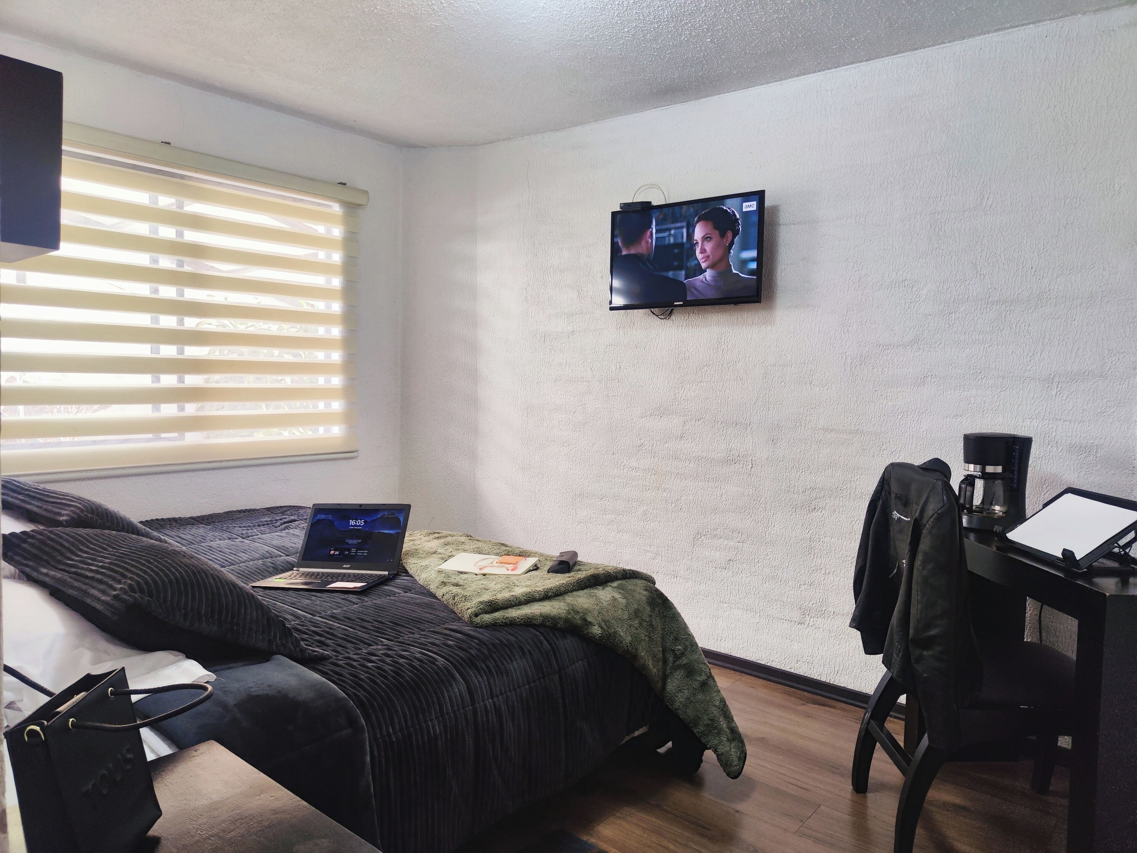 A small, neatly arranged bedroom with a bed, a desk, a coffee maker, and a TV on the wall.