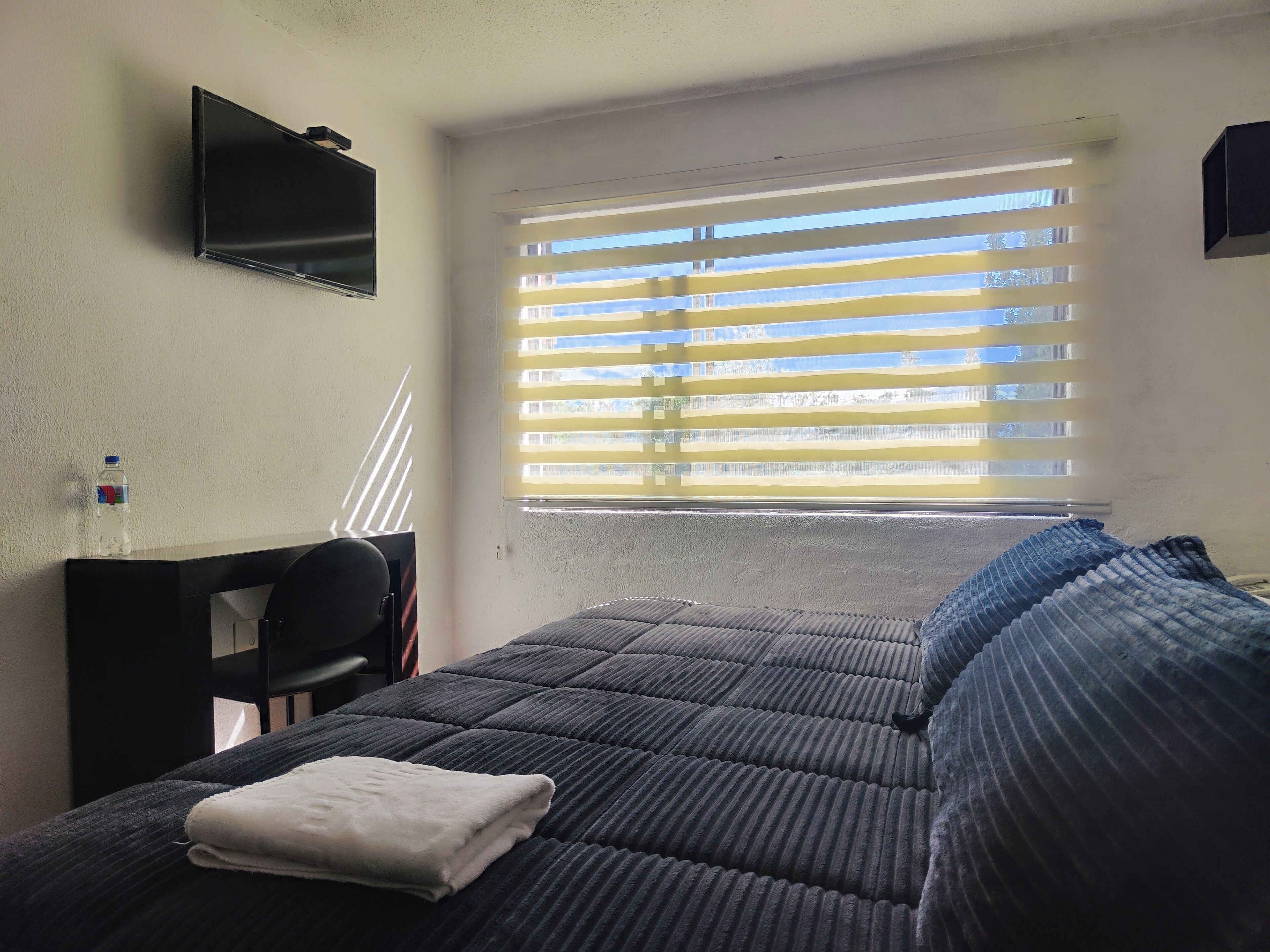 A hotel room with a bed, a desk, a chair, a TV mounted on the wall, and a window with blinds.
