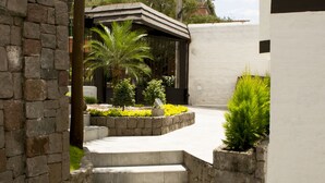Un sendero conduce a un edificio con un muro de piedra a un lado y un muro blanco al otro. Hay escalones que suben al edificio, y un pequeño jardín con plantas y flores.