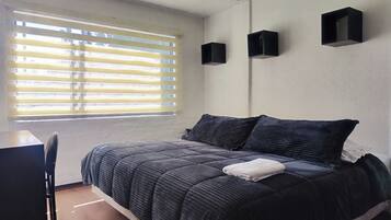 A bedroom with a bed, a window with blinds, a desk, and two wall-mounted speakers.