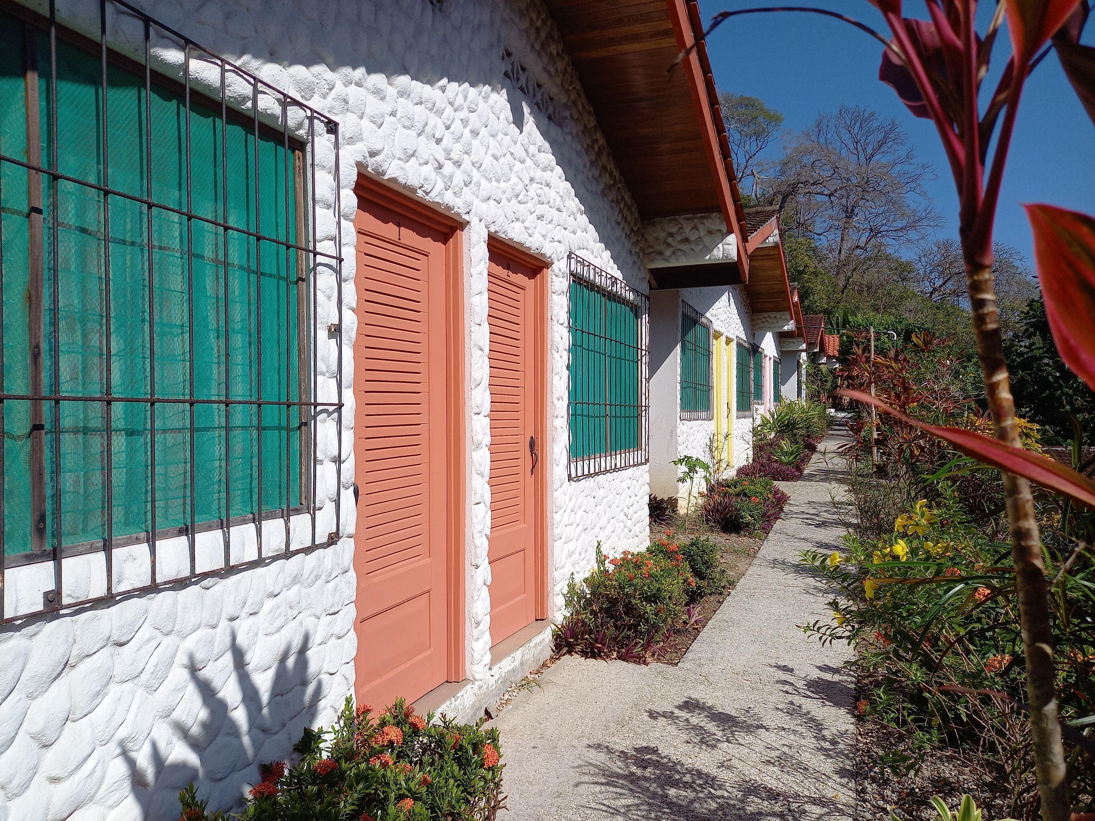 Una hilera de edificios blancos con contraventanas verdes y puertas rojas, bordeada de plantas coloridas bajo un cielo despejado.