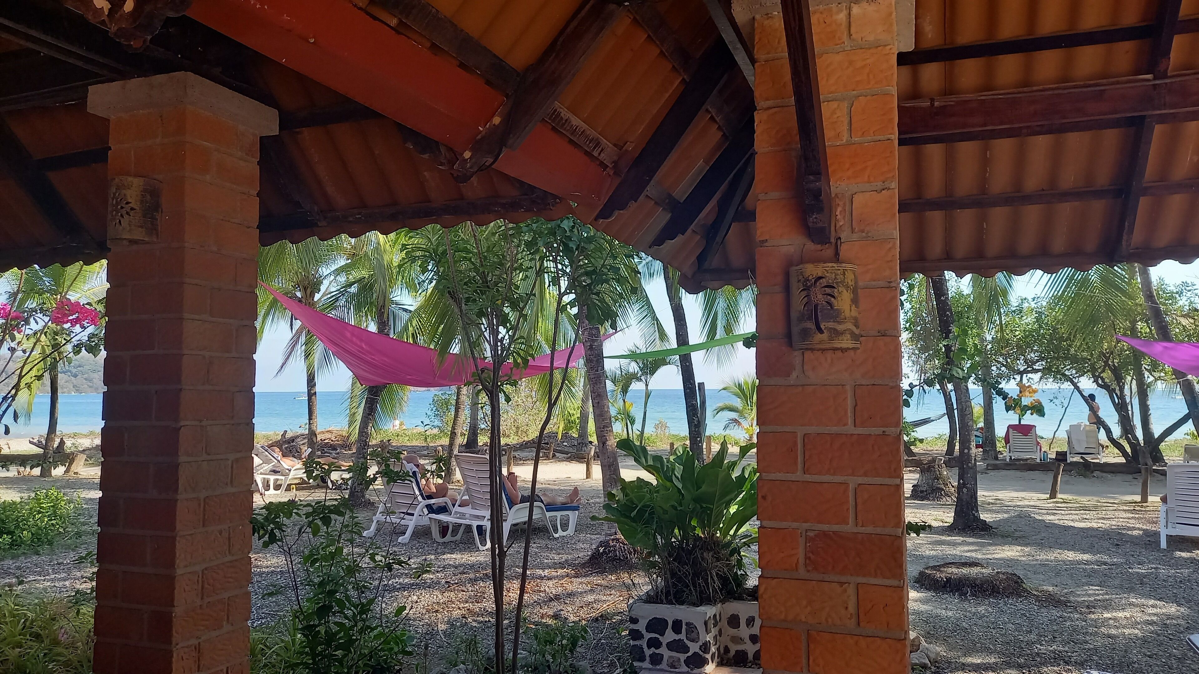 A covered outdoor seating area with brick pillars, a view of the beach, and palm trees.