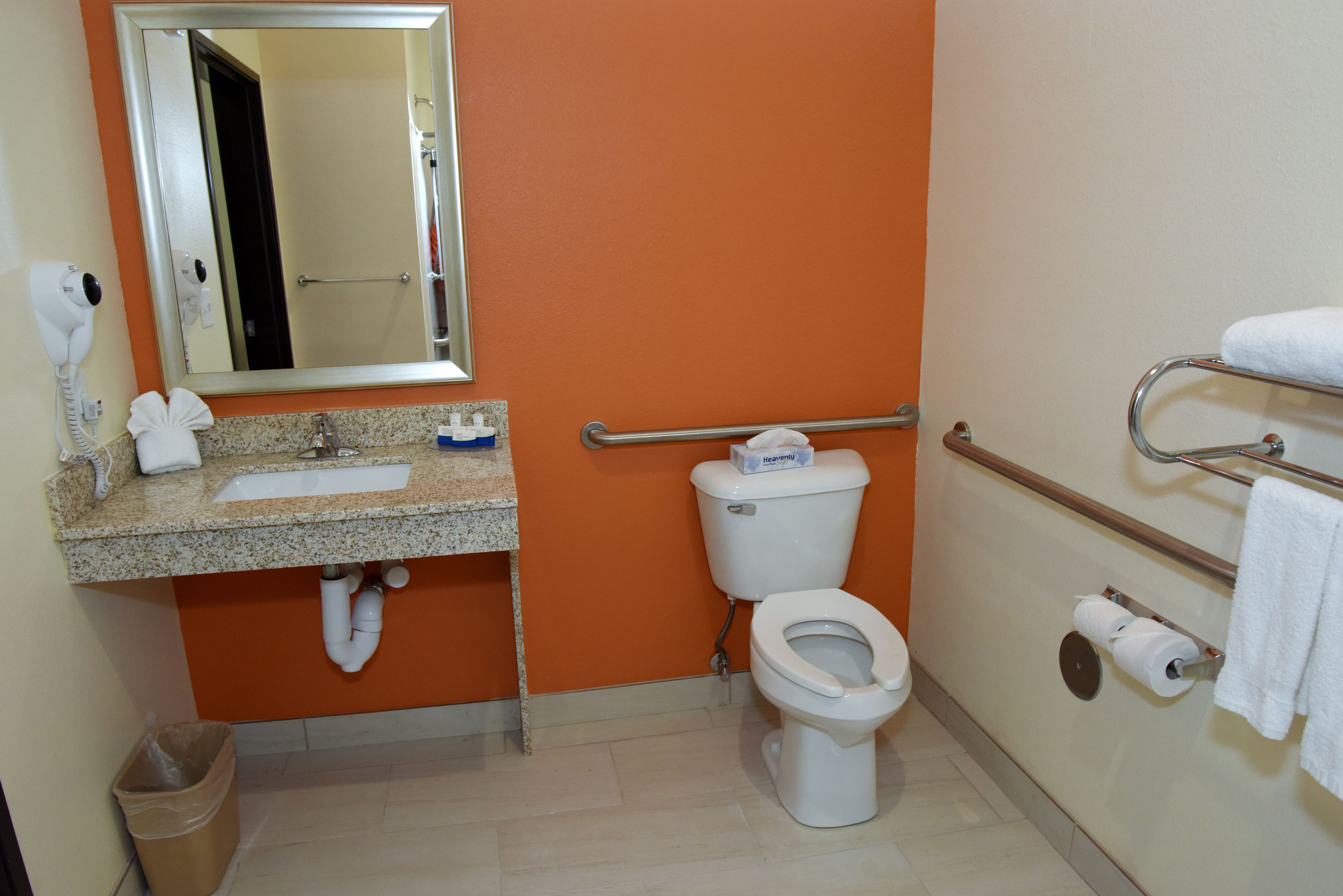 A bathroom with an orange wall, a sink with a mirror above it, a toilet, a trash can, and a towel rack with towels.
