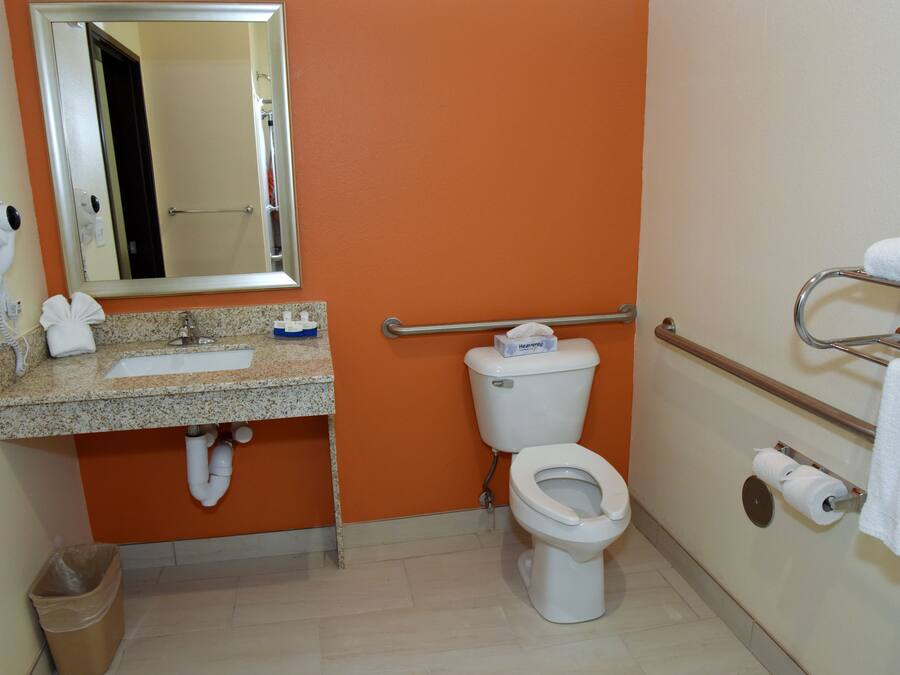 A bathroom with an orange wall, a sink with a mirror above it, a toilet, a trash can, and a towel rack with towels.