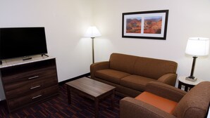 A hotel room with a flat-screen TV on a wooden cabinet, a brown sofa, an orange armchair, a small wooden coffee table, and two floor lamps.