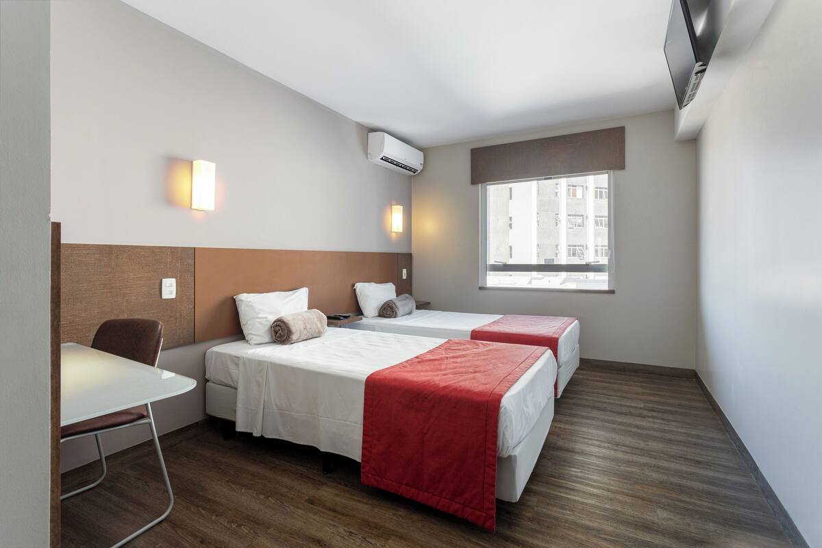 Ein Hotelzimmer mit zwei Betten, einem Schreibtisch, einem Sessel und einem Fenster mit Blick auf Gebäude. Eine Klimaanlage ist ebenfalls vorhanden.