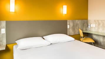 Habitación de hotel con cama, dos almohadas, una silla y dos lámparas fijadas a la pared.