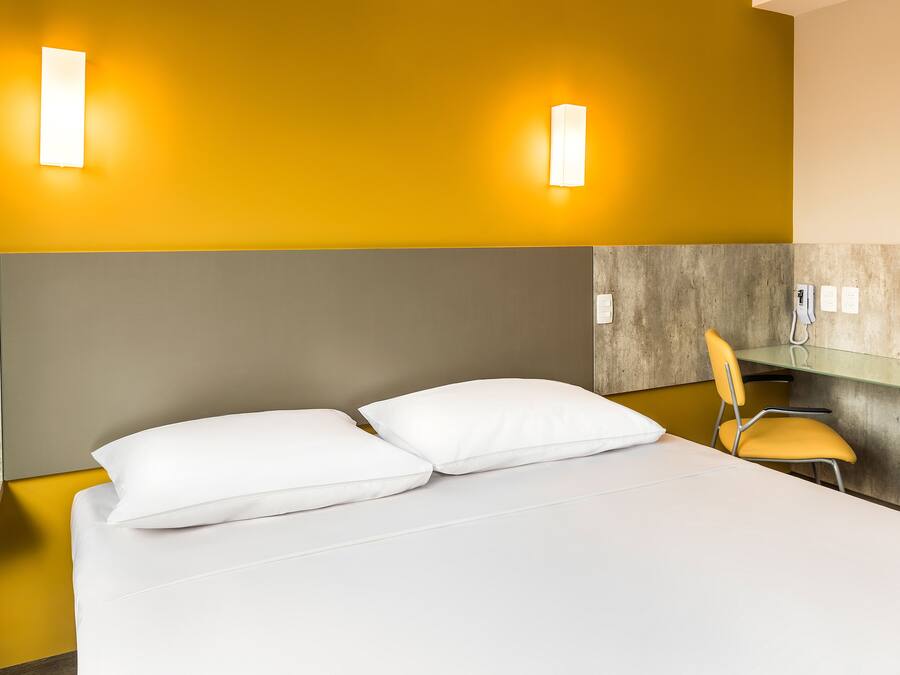 Habitación de hotel con cama, dos almohadas, una silla y dos lámparas fijadas a la pared.
