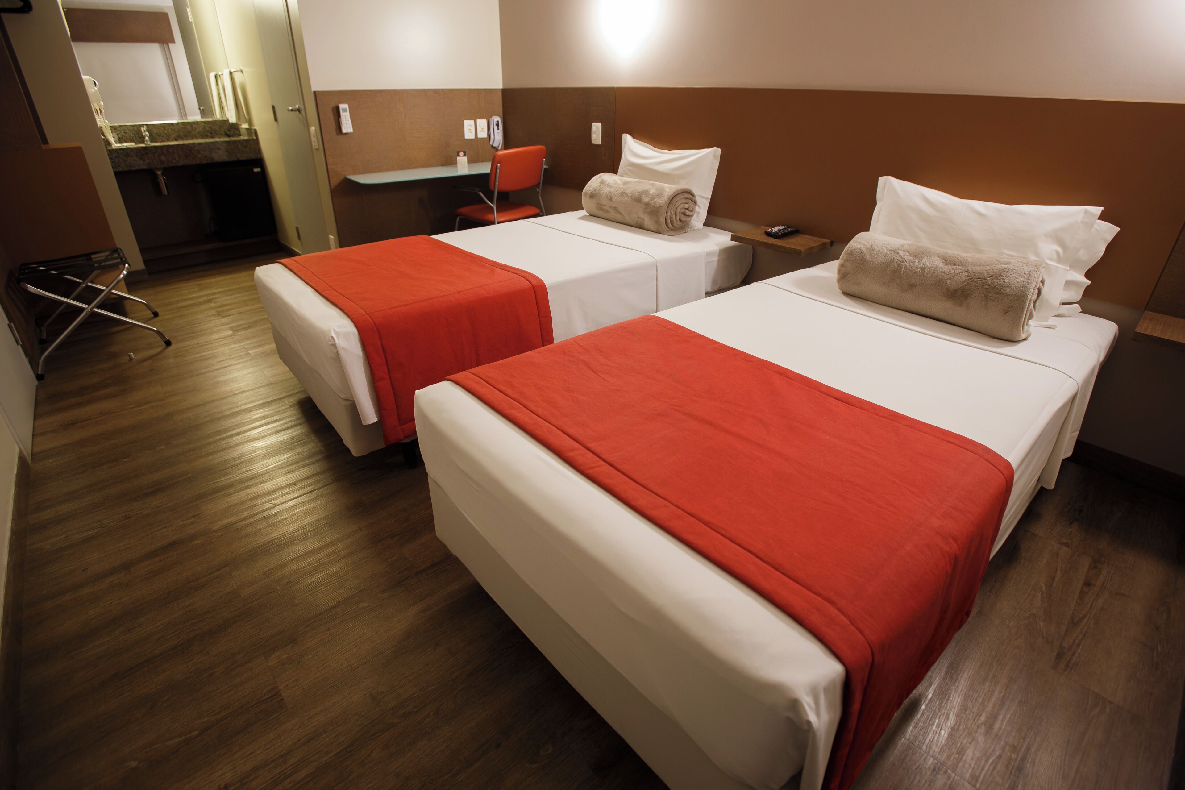 A hotel room with two beds, each with a red bedspread and white pillows, a small desk with a chair, and a bathroom visible in the background.