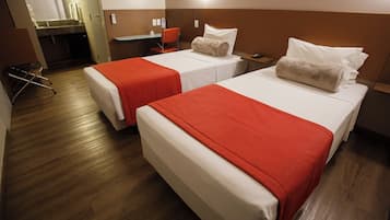 Habitación de hotel con dos camas, cada una con una colcha roja y almohadas blancas, un escritorio pequeño con una silla, y un baño visible al fondo.