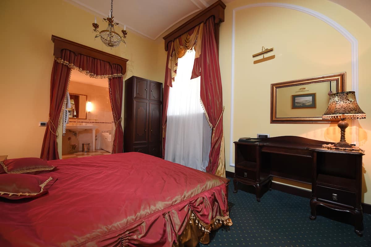 A bedroom with a large bed, a desk, a chair, and a dresser. There is a window with curtains and a chandelier.