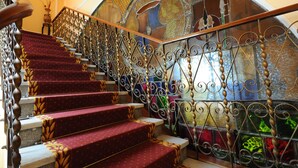 Un magnifique escalier, orné de rampes en métal ouvragé et de marches recouvertes de moquette rouge, mène à une vaste fenêtre en vitrail en forme d’arc.