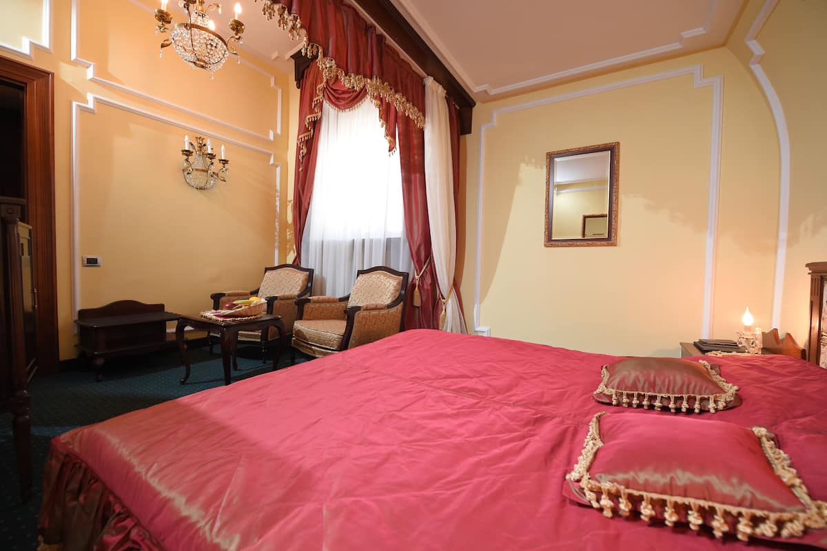A hotel room with a large bed, two armchairs, a small table, a chandelier, and a mirror on the wall.