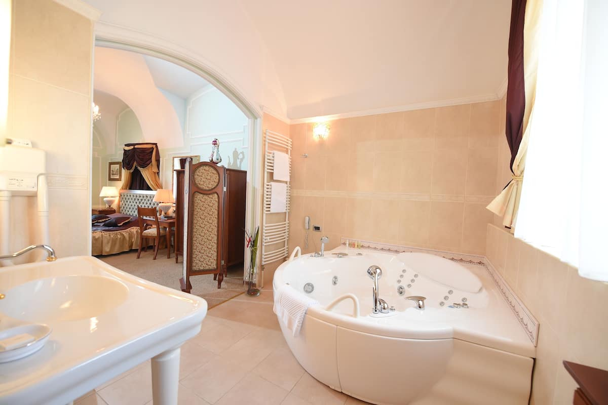 A bathroom with a large bathtub, a sink, and a vanity. There is a mirror above the sink and a towel rack beside it. The room has a tiled floor and a window with curtains.